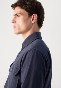 Mørkeblå skjorte med knapper og krage, med en brystlomme og en jevn tekstur, vist på en modell fra en sideprofil.