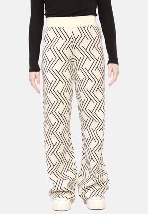 Persona che indossa pantaloni bianchi con linee a zigzag nere, scarpe bianche e una maglia nera a maniche lunghe, in piedi contro uno sfondo semplice.