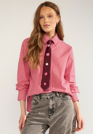 Femme aux longs cheveux portant une chemise rayée rouge et blanche, une cravate rouge foncé avec des décorations florales, et un jean gris délavé.