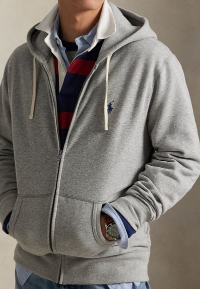 Mann trägt geschichtete blaue Hemden mit Kragen und Streifen unter einem grauen Zip-Hoodie, mit den Händen in den Taschen und einer sichtbaren Armbanduhr.