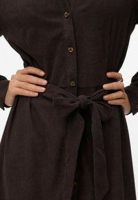 Tumepruun sametist kleit, millel on esiküljel nööbid, vöökohal lipsuga vöö ja pikad varrukad, rõhutades soonilist tekstuuri ja klassikalist disaini.