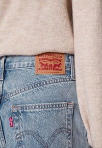 Ljusblå denimjeans med en beige läderlapp som har röda grafiska tryck och text. Tyget är strukturerat med synliga sömmar och en fickdetalj.