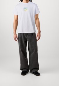 Valkoinen puuvillainen t-paita, jossa värikäs kuvio rinnassa, yhdistetty löysiin tummanharmaisiin farkkuihin ja mustiin kenkiin.