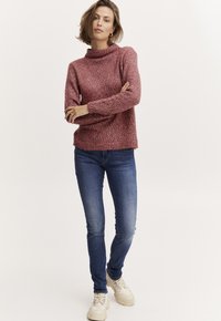 Adīts bordo džemperis ar augstu apkakli, garām piedurknēm un ripotā struktūrā, apvienots ar ziliem džinsiem un bēšu sporta apaviem.