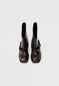 Stivaletti alla caviglia in pelle marrone scuro con punta quadrata, fascia decorativa e fibbia dorata, mostrati frontalmente su uno sfondo neutro.