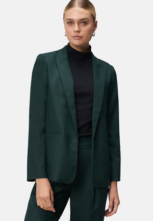 Femme aux cheveux blonds portant des boucles d'oreilles en or, un blazer vert foncé, un pantalon assorti et un col roulé noir, se tenant devant un fond clair.