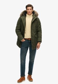 Parka verde con capucha forrada de piel, suéter de punto trenzado beige, jeans azules y botas de cordones marrones. Presenta bolsillos y un diseño informal.