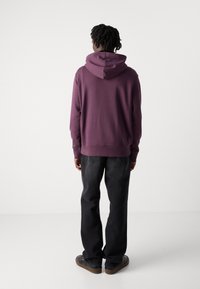 Kapuzensweatshirt in tiefem Lila, aus weichem Stoff gefertigt. Mit gerippten Bündchen und Saum, getragen mit schwarzen Jeans und lässigem Schuhwerk.