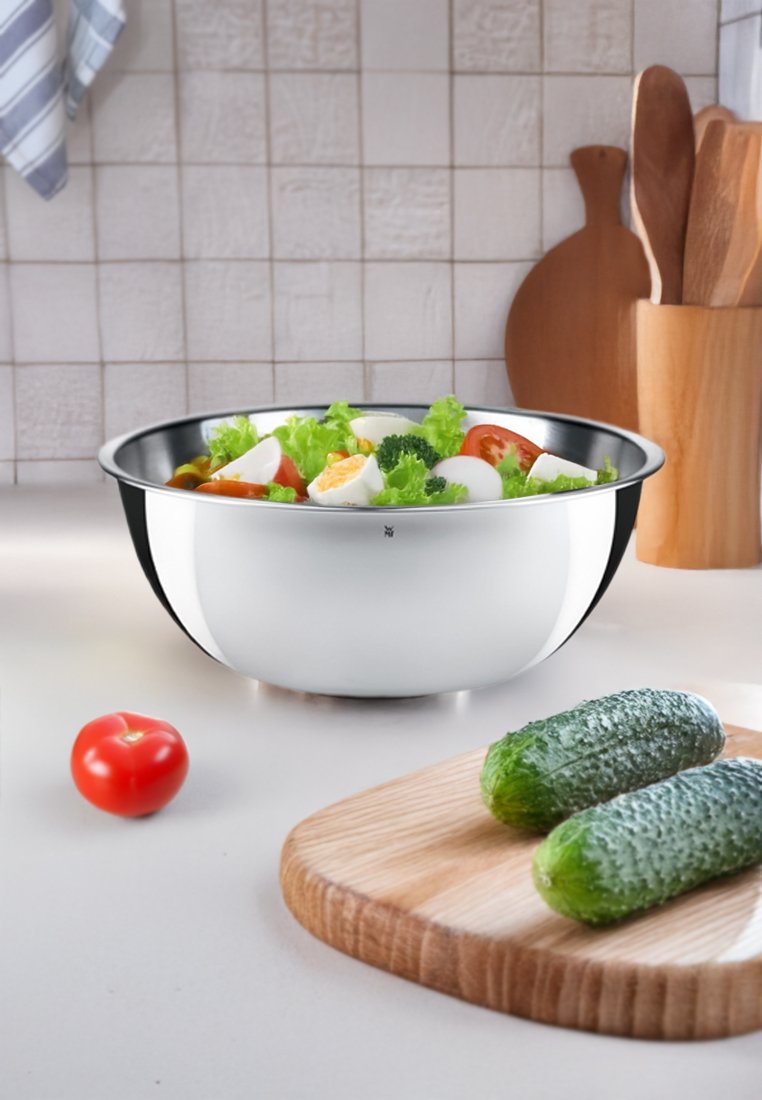 Ensaladera de acero inoxidable con un acabado pulido, llena de lechuga, tomates cherry, huevos y brócoli. Tabla de cortar de madera con pepinos.
