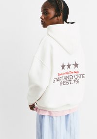 Overgrote witte hoodie met een trekkoordkap, voorzien van een grafische print met sterren en tekst op de achterkant. Zachte, fleecachtige textuur.