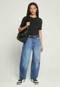 Mulher jovem com cabelo longo e encaracolado, vestindo uma blusa preta de mangas curtas, jeans azul largo e rasgado, sapatilhas preto e branco, e a segurar uma grande mala preta de ombro.