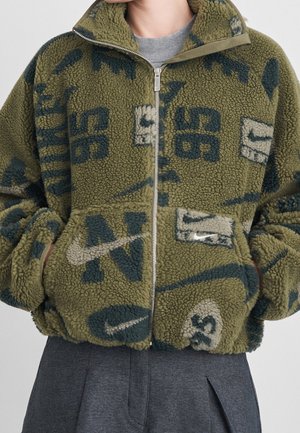 Personne portant une veste polaire verte avec des chiffres et des logos graphiques bleus et beiges, fermée par une fermeture éclair, mains dans les poches, et pantalon gris.