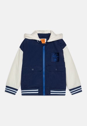 Veste bleu marine avec une capuche blanche, fermeture éclair, deux poches avant, poignets rayés et un logo sur patch. Fabriquée en tissu léger.