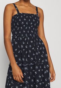 Blommig klänning med marinblå bakgrund. Har smockad byst, tunna axelband och en flerlagerskjol med små blå och gröna blommönster.