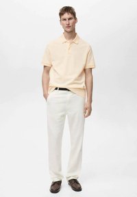 Světle oranžové polo tričko s límečkem v kombinaci s bílými kalhotami a hnědými botami, stojící na jednobarevném pozadí.