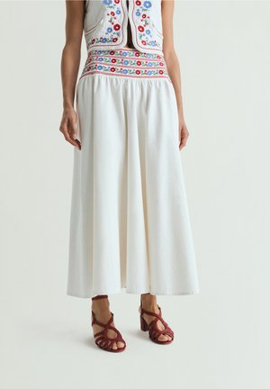 Mujer con falda larga blanca con bordado floral en la cintura, combinada con un chaleco bordado con flores y sandalias rojas de tacón.