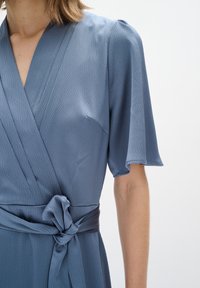 Blaues Wickelkleid aus strukturiertem Stoff, mit kurzen Ärmeln, V-Ausschnitt und gebundenem Taillenbereich. Glatte Oberfläche mit einem dezenten Muster.