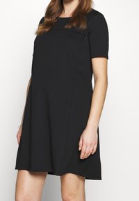 Donna in un vestito nero a maniche corte che sta in piedi contro uno sfondo bianco semplice, con capelli castani sciolti e un braccialetto al polso sinistro.