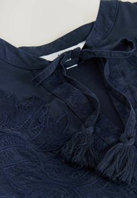 Námořnická modrá blůza s detailním vyšíváním listů, stahovací šňůrkou s třásněmi a kulatým výstřihem. Hladká tkanina s jemnou texturou.