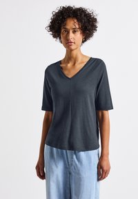 Koszulka w kolorze węglo-szarym z dekoltem w serek, wykonana z miękkiego materiału. Krótkie rękawy, luźny krój oraz subtelne detale szwów. Połączona z jasnoniebieskimi dżinsami.