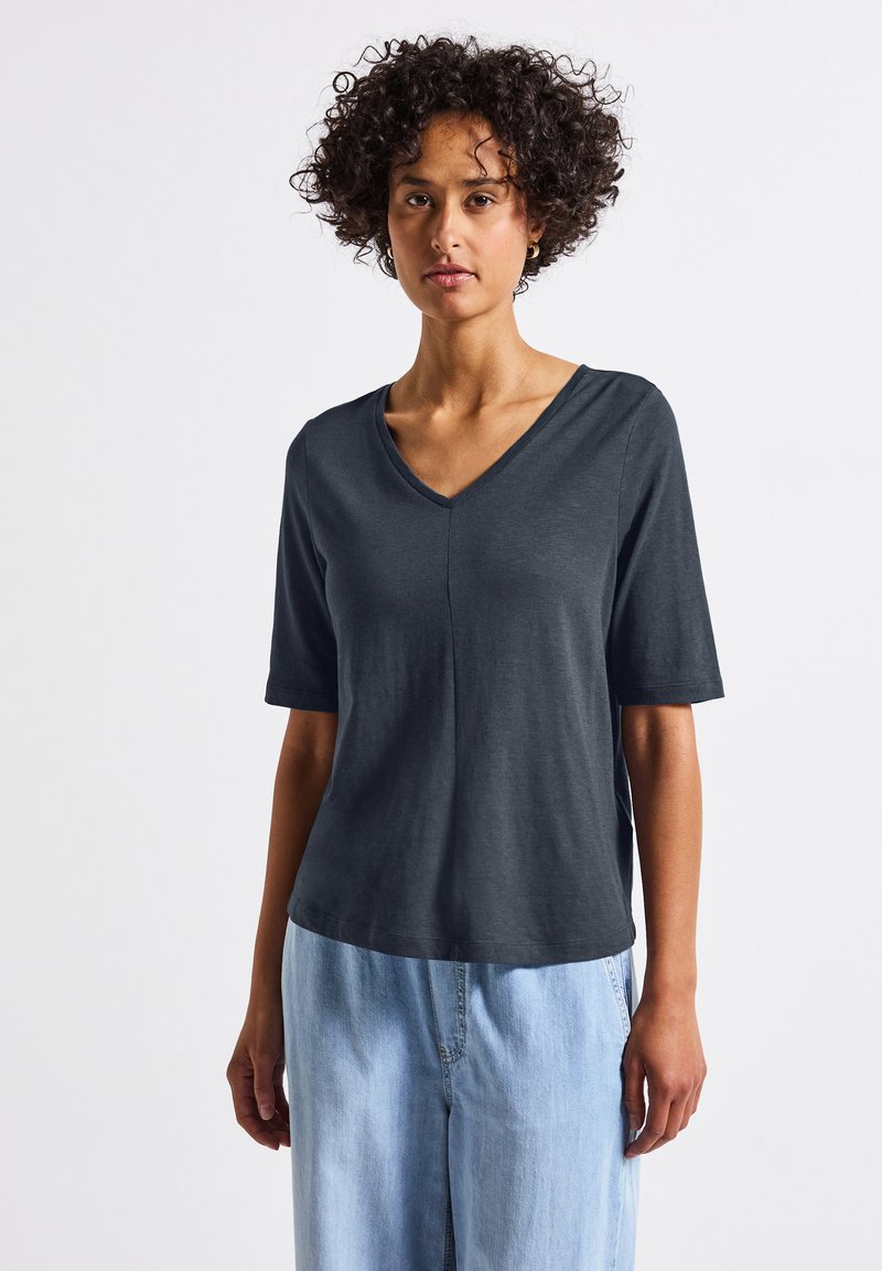 Koszulka w kolorze węglo-szarym z dekoltem w serek, wykonana z miękkiego materiału. Krótkie rękawy, luźny krój oraz subtelne detale szwów. Połączona z jasnoniebieskimi dżinsami.