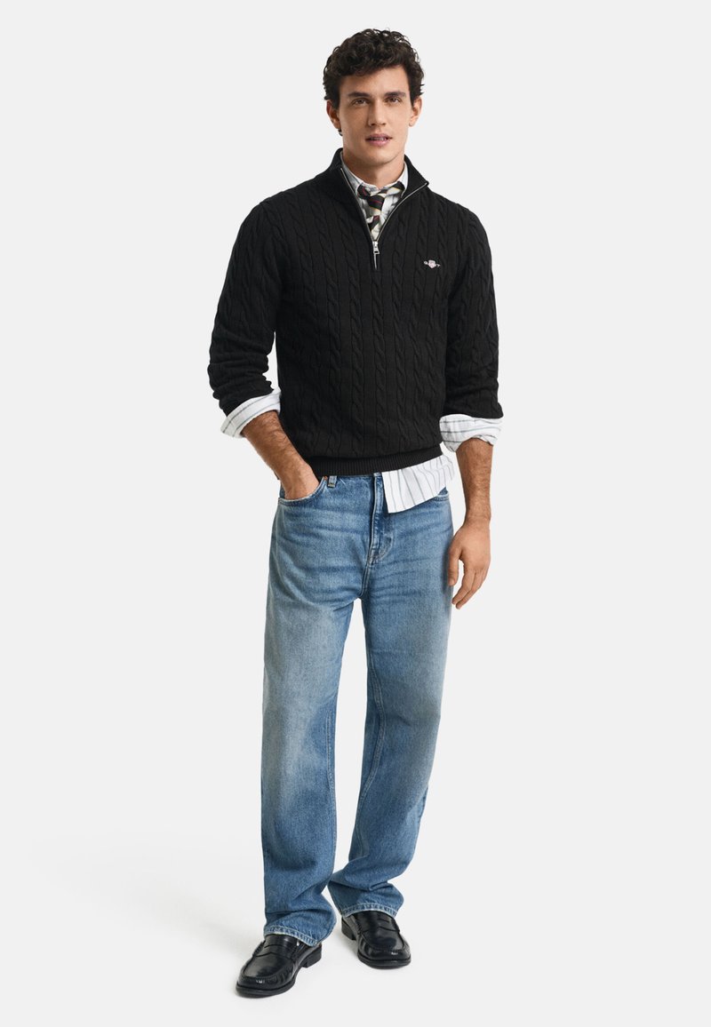 Man wearing black cable-knit quarter-zip sweater over striped shirt with rolled sleeves, light blue jeans, and black shoes, standing casually.