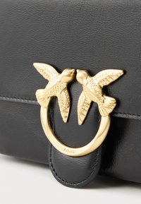 Fecho de uma bolsa de couro preto com duas aves douradas detalhadas a enfrentar-se, ligadas por um anel dourado abaixo dos seus bicos.