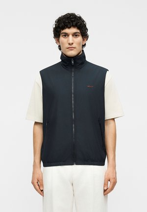 Άνδρας με σγουρά μαλλιά που φοράει μαύρο αμάνικο γιλέκο με φερμουάρ πάνω από ένα λευκό κοντομάνικο πουκάμισο, στέκεται μπροστά σε ένα απλό φωτεινό φόντο.