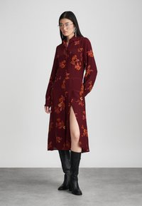 Μπορντό φόρεμα με λουλούδια, μακριά μανίκια, μπροστινό κλείσιμο με κουμπιά και πλαϊνό σκίσιμο. Κατασκευασμένο από απαλό ύφασμα με κίτρινες και πορτοκαλί λουλουδένιες λεπτομέρειες.