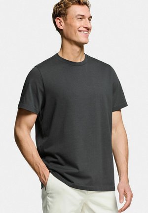 Glunderende man die een effen zwart T-shirt met korte mouwen en witte broek draagt, met één hand in zijn zak, staand tegen een witte achtergrond.