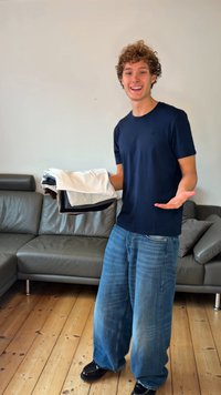 Donkerblauw T-shirt, losse pasvorm, gecombineerd met brede lichtblauwe jeans. Model houdt een stapel opgevouwen shirts vast in wit, grijs en zwart.