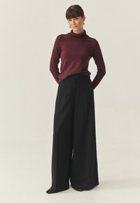 Burgundy garbós pulóver széles lábú fekete nadrággal párosítva. Sima textúra, szabott fazon és minimalista dizájn hegyes fekete cipőkkel.