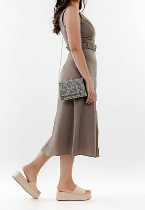 Žena v bezramenných taupe šatech s páskem, béžových platformových pantoflích a nese malou černobílou kostkovanou kabelku přes rameno.