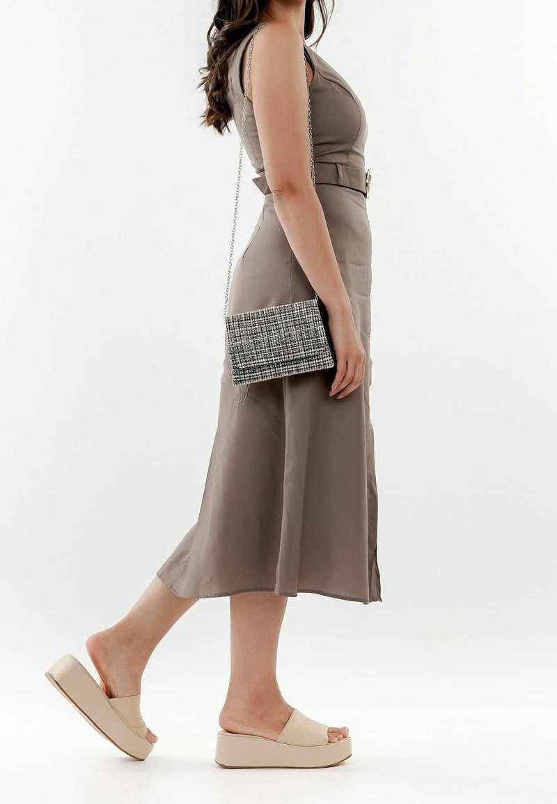 Kobieta ubrana w bezrękawnik w odcieniu taupe, z paskiem, beżowe sandały na platformie oraz nosząca małą torebkę na krzyż w czarno-białą kratkę.