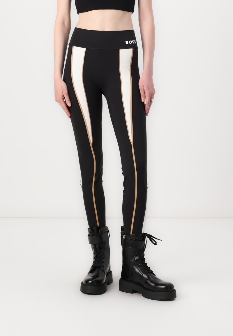 Fekete leggings fehér és arany oldalsávokkal, magas derékkal, fekete fűzős katonai csizmákkal, vastag talppal és cipzáras díszítéssel párosítva.