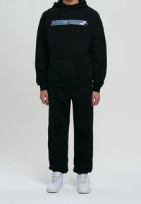 Zwarte hoodie met "LOWLIGHTS" grafisch in blauw aan de voorkant. Gecombineerd met zwarte joggingbroek en witte sneakers. Zachte, casual stof.