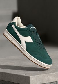 Green suede sneaker with white accents, a textured rubber sole, and lace-up design. Features a logo on the tongue and side.