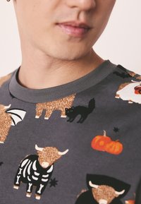 Sweatshirt gris avec différents motifs d'animaux, dont un chat noir, une citrouille et des vaches dessinées de manière cartoon. Encolure côtelée. Texture de tissu douce.