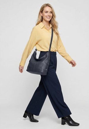 Lachende vrouw die loopt, gekleed in een gele blouse, marineblauwe brede broekspijpen, zwarte laarzen met hakken, en een grote navy leren schoudertas draagt.