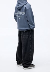 Sudadera gris con texto blanco en la parte posterior, combinada con jeans negros holgados. El calzado incluye zapatillas deportivas grises. Diseño simple y casual.