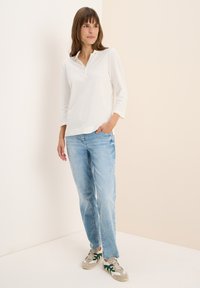 Witte longsleeve polo met een knoopsluiting, gecombineerd met lichtblauwe jeans en beige sneakers met groene en grijze accenten.