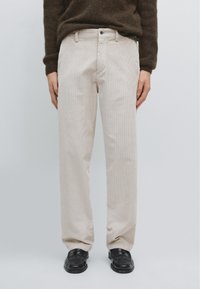 Pantalon en velours côtelé beige avec une coupe droite, doté d'une fermeture à bouton et de poches avant, assorti de chaussures noires.
