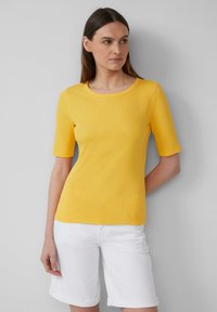 Femme portant un haut jaune à manches courtes et un short blanc arrivant aux genoux, se tenant devant un fond gris clair uni.