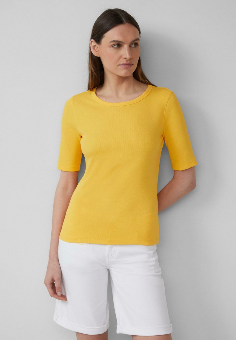 Femme portant un haut jaune à manches courtes et un short blanc arrivant aux genoux, se tenant devant un fond gris clair uni.