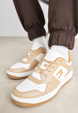 Beżowe i białe sneakersy z etykietą "Tommy Jeans", noszone z białymi skarpetkami i brązowymi spodniami z podwiniętymi nogawkami na jasnej powierzchni podłogi.