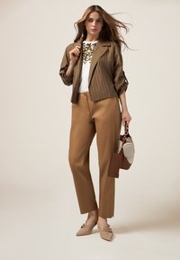 Blazer a righe marrone sopra una maglietta bianca con decorazioni, pantaloni color tan e scarpe beige a punta; tiene in mano una borsa multicolore.