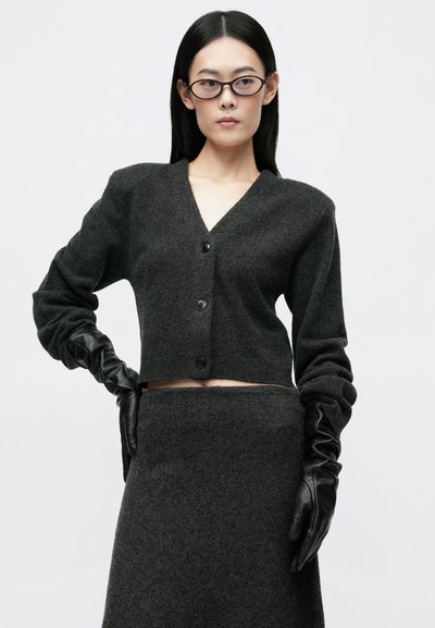 Καρφίτσα σε σκούρο γκρι χρώμα, σε μαλακό ύφασμα πλέξης, με V-λαιμόκοψη, κλείσιμο με κουμπιά και μακριά μανίκια. Συνδυάζεται με μια ταιριαστή γκρι φούστα και μαύρα δερμάτινα γάντια.