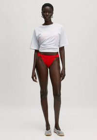 Weißes, verkürztes T-Shirt, roter Bikini-Unterteil mit seitlichen Knotendetails, hellgraue Ballerinas, Model mit kurzem schwarzen Haar und Creolen.