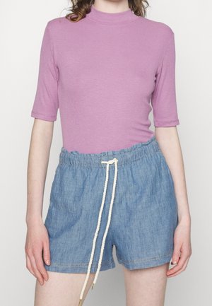 Person trägt ein fliederfarbenes kurzärmeliges Oberteil, das in hellblaue hoch taillierte Jeansshorts mit weißem Kordelzug gesteckt ist.