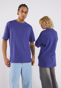 Violet kokvilnas T-krekli ar īsām piedurknēm, brīva katura, ar sānu šķēlumiem, ko valkā vīrietis un sieviete, attiecīgi pieskaņotiem gaišiem un tumšiem džinsiem.